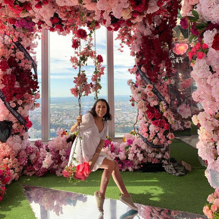 Mariela sitting on a swing surrounded by columns of roses with high rise window overlooking city