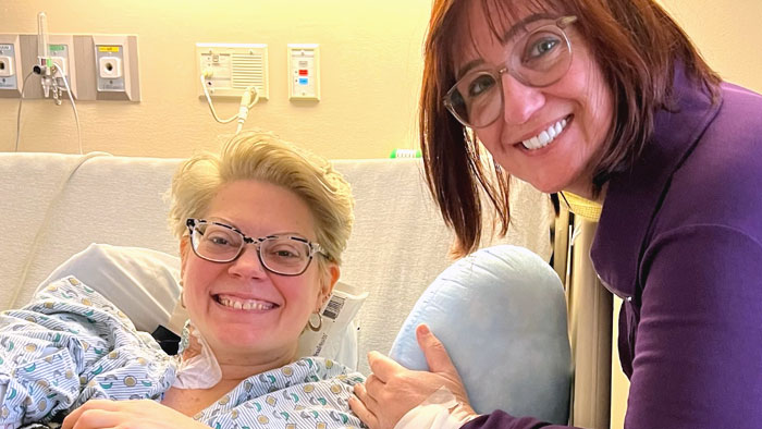 Two women in hospital room. One sitting on hospital bed, the other leaning over, both smiling