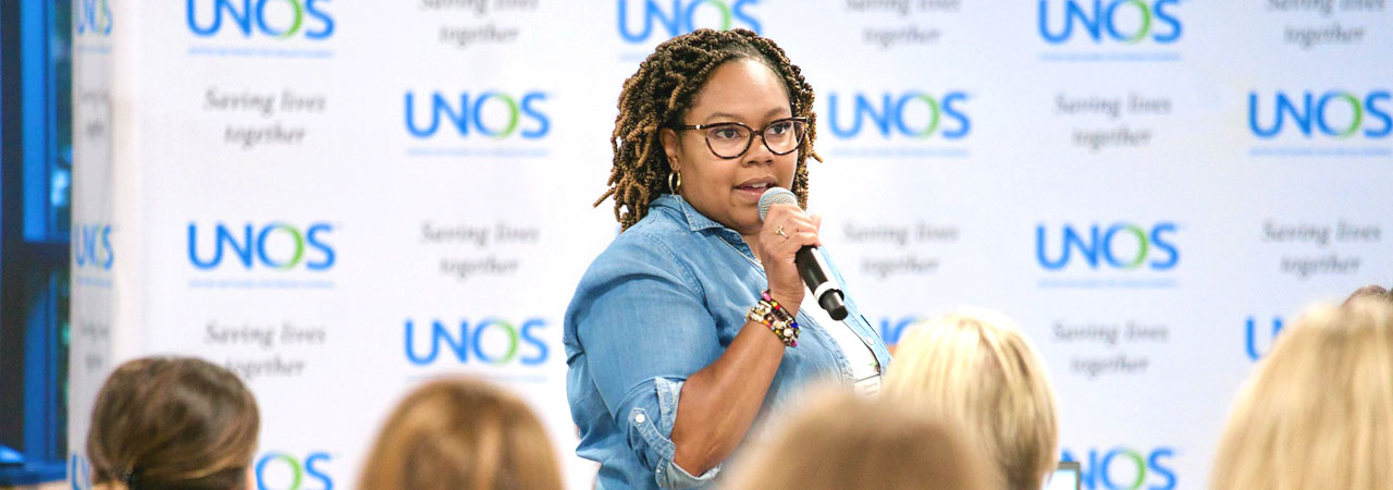 Woman in red jacket holding microphone to address participants at 2023 UNOS Primer