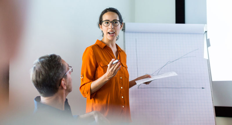 Woman holding papers standing in front of a large presentation notebook, talking